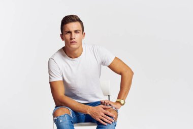 Sandalyede oturan adam yırtık kot pantolonlu, beyaz tişörtlü yakışıklı, açık renk arka planlı.