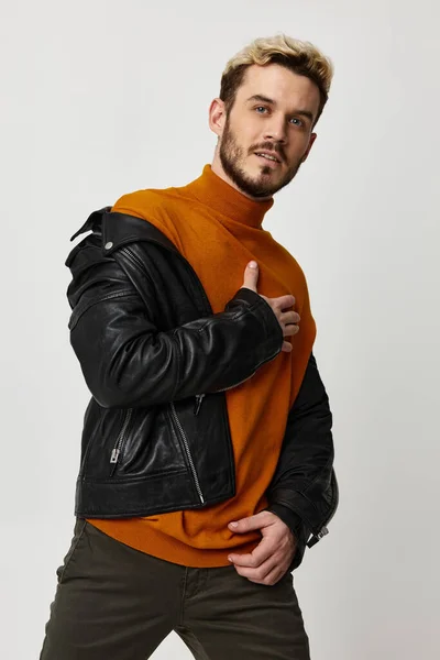 Блондин в кожаной куртке с оранжевым свитером и брюками позирует на светлом фоне — стоковое фото