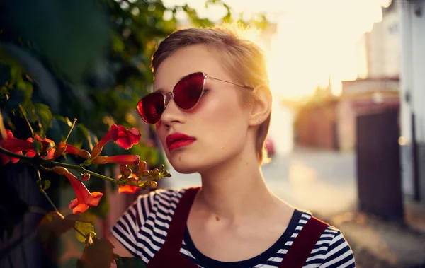 Vacker kvinna i glasögon nära röda blommor på gatan och röd solklänning randig t-shirt — Stockfoto