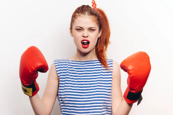 Mujer pelirroja con guantes de boxeo y una camiseta a rayas sobre un fondo claro con la boca abierta —  Fotos de Stock