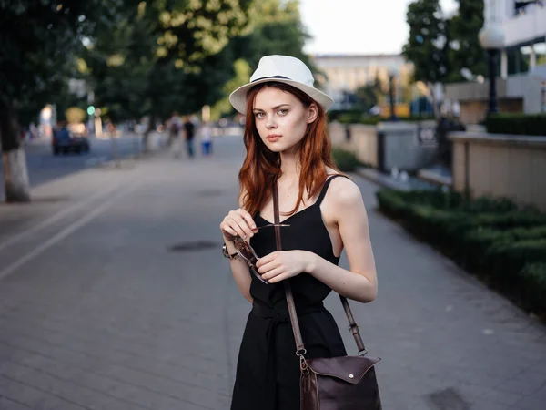 Een mooie vrouw in een jurk en hoed met een tas op haar schouder loopt door de straat — Stockfoto