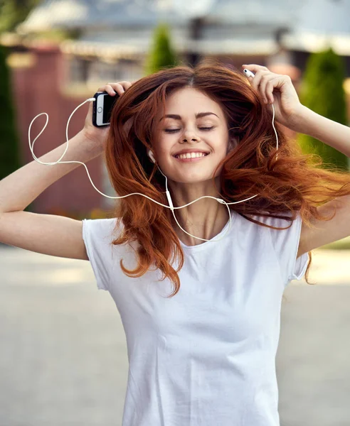 Mulher em uma camiseta branca caminha pela rua em fones de ouvido com um telefone — Fotografia de Stock