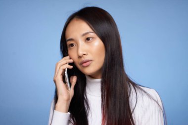 Asyalı kadın mavi arka planda cep telefonuyla konuşuyor.