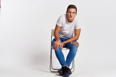 Sandalyede oturan adam yırtık kot pantolonlu, beyaz tişörtlü yakışıklı, açık renk arka planlı.