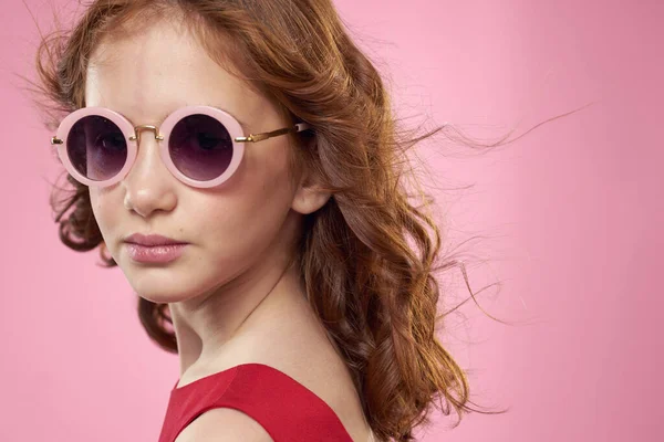 Дівчина з кучерявим волоссям темні круглі окуляри весела червона сукня рожевий фон — стокове фото