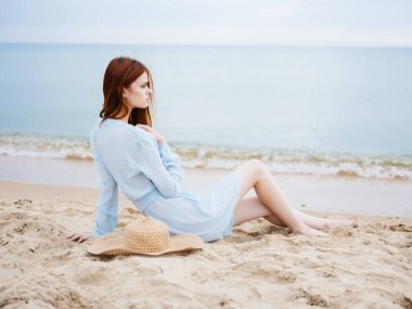 Denize yakın bir elbisenin içinde elinde şapkayla kumların üzerinde duran kadınlar.