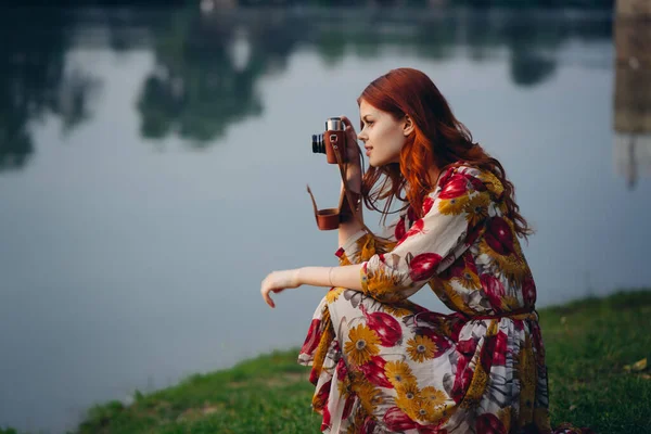Κοκκινομάλλα γυναίκα με μια φωτογραφική μηχανή στη φύση κοντά στη λίμνη — Φωτογραφία Αρχείου