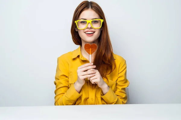 Aan tafel zit een vrolijke vrouw met een gele bril en een hartvormige lolly — Stockfoto