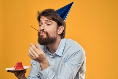 Elinde doğum günü pastası ve doğum gününde sarı arka planda şapkası olan bir adam.