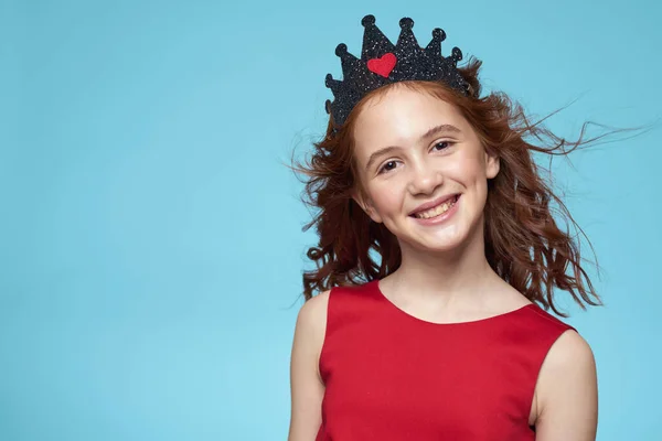 Holčička s kudrnaté vlasy s korunou na hlavě červené šaty úsměv modré pozadí — Stock fotografie