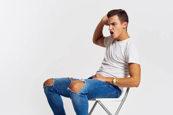 Man op een stoel binnen gescheurde jeans wit t-shirt knappe gezicht model lichte achtergrond — Stockfoto