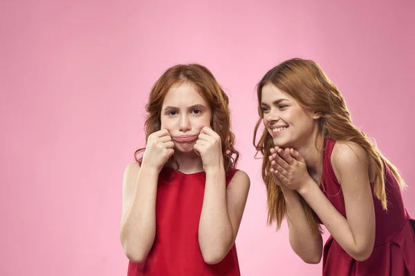 Mamá e hija en vestidos rojos abrazos divertido mueca infancia alegría rosa fondo — Foto de Stock