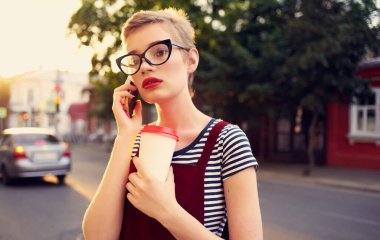 Elinde bir fincan kahveyle sokakta telefonla konuşan ve gözlüklü bir sarışın.