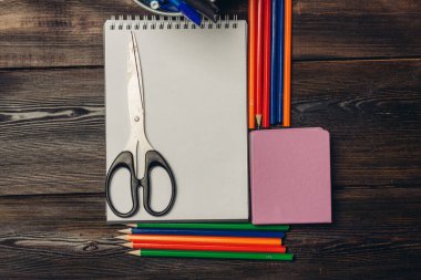 Makas açık bir deftere, renkli kalemler de ahşap bir masa görünümüne uzanır.