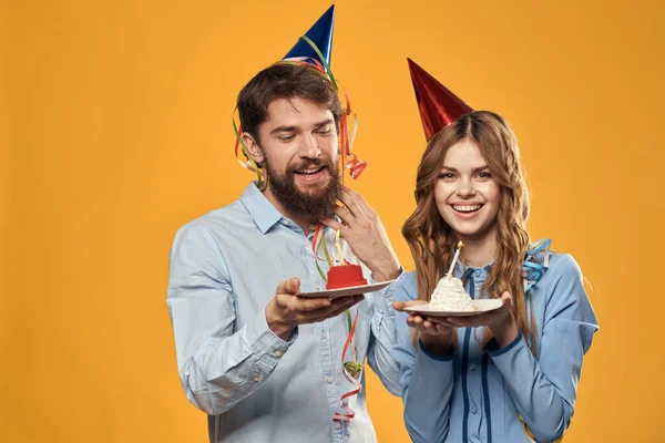 Energický muž a hezká žena s narozeninovým dortem v klobouky na žlutém pozadí disco party — Stock fotografie