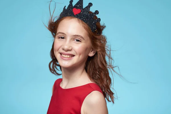 Bambina con i capelli ricci con una corona sulla testa vestito rosso Sorriso sfondo blu — Foto Stock