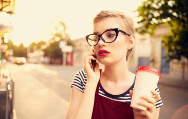Elinde bir fincan kahveyle sokakta telefonla konuşan ve gözlüklü bir sarışın.
