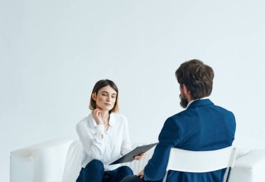 Elinde belgeler olan bir kadın psikolog koltukta oturuyor ve takım elbiseli bir adam da sandalyede.