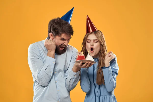 Aniversário festa homem e mulher em um fundo amarelo em chapéus com um bolo em suas mãos — Fotografia de Stock