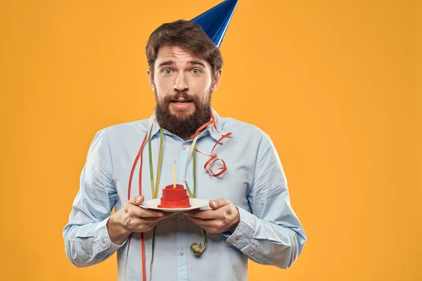 Hombre alegre con un pastel en un fondo amarillo cumpleaños días festivos gorra en su cabeza — Foto de Stock