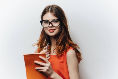 Portakal rengi bir elbise giymiş güzel bir öğrenci. Elinde bir not defteri var. Hafif bir arkaplan üzerinde el hareketi yapıyor.