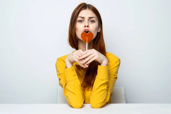 Hübsche Frau sitzt am Tisch mit herzförmigem Lutscher gelbes Hemd — Stockfoto