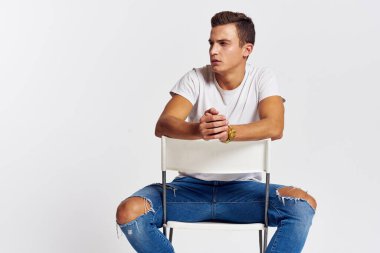 Sandalyede oturan adam yırtık kot pantolonlu, beyaz tişörtlü yakışıklı, açık renk arka planlı.