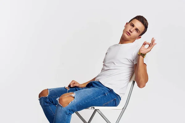 Man op een stoel binnen gescheurde jeans wit t-shirt knappe gezicht model lichte achtergrond — Stockfoto