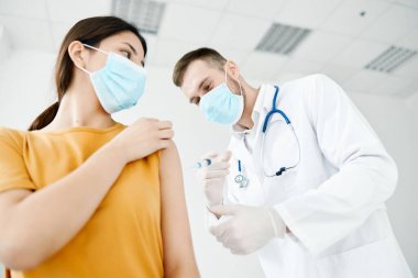 Doktor, tıbbi maske takan bir hastanın omzuna iğne yapıyor. Alttan görüşlü kovan aşısı.