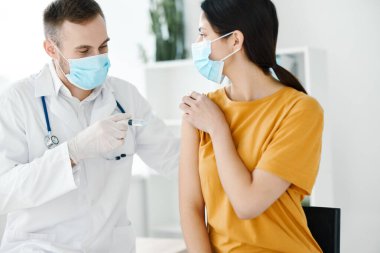 Doktor, hastane kovid aşısıyla sarı tişörtlü bir kadının omzuna iğne yapıyor.