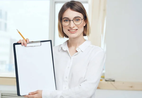 Eine Frau hält einen Ordner mit einem weißen Blatt Papier und einem Fenster im Hintergrund — Stockfoto