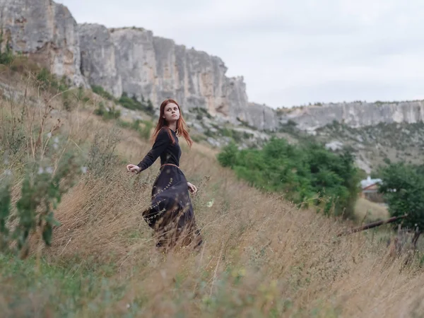 Mujer viajero en un vestido negro corre sobre hierba seca en el prado y las montañas en el fondo —  Fotos de Stock