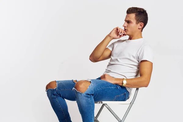 Uomo su una sedia indoor strappato jeans bianco t-shirt bel viso modello luce sfondo — Foto Stock