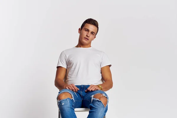 Hombre en una silla interior desgarrado jeans blanco camiseta guapo cara modelo luz fondo — Foto de Stock