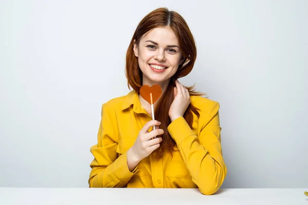 Mulher atraente alegre com um pirulito em forma de coração se senta à mesa — Fotografia de Stock