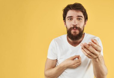 Beyaz tişörtlü, sarı arka planda elleri kırpık bir şekilde el kol hareketi yapan duygusal adam.