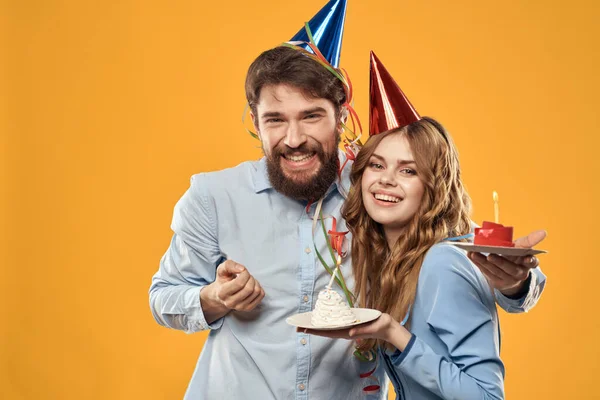 Homem enérgico e mulher bonita com bolo de aniversário em chapéus em uma festa disco fundo amarelo — Fotografia de Stock