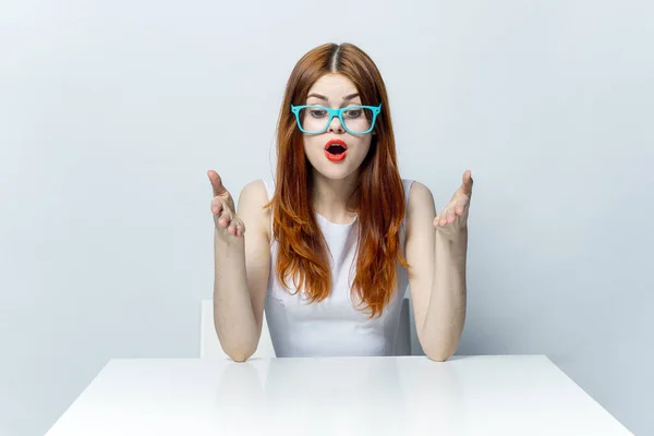 Mooie roodharige vrouw zitten aan de tafel met een blauwe bril rode lippen emoties glamour — Stockfoto