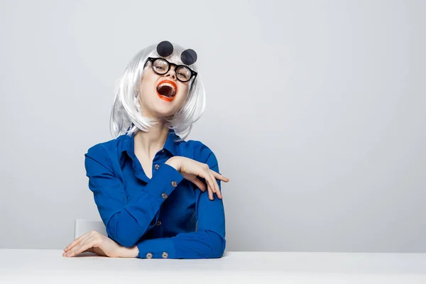 Vrolijke vrouw in een witte pruik rode lippen donkere bril zit aan een tafel in een blauw shirt — Stockfoto