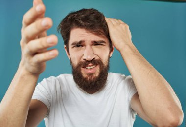 Sakallı üzgün bir adam mavi arka planda elleri ile kafasına dokunur.