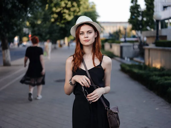 Una mujer bonita con un vestido y un sombrero con una bolsa en el hombro camina por la calle —  Fotos de Stock