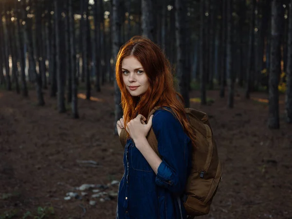 Červenovlasá žena v modré košili v borovém lese s batohem na zádech — Stock fotografie