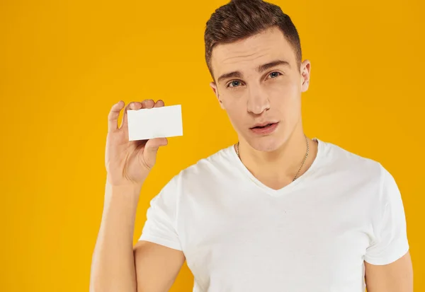 Een man in een wit t-shirt toont een creditcard op een gele achtergrond bijgesneden uitzicht — Stockfoto