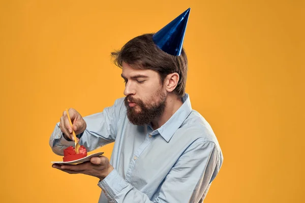 Elinde doğum günü pastası ve doğum gününde sarı arka planda şapkası olan bir adam. — Stok fotoğraf