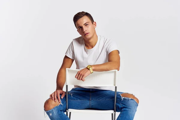 Muž na židli uvnitř roztržené džíny bílá tričko hezký obličej model světlo pozadí — Stock fotografie