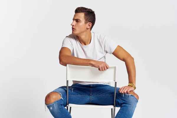 男人在屋里的椅子上撕破牛仔裤白色T恤英俊的脸型浅色背景 — 图库照片