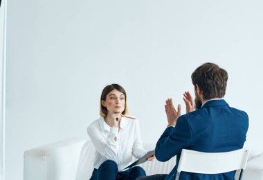 Elinde belgeler olan bir kadın psikolog koltukta oturuyor ve takım elbiseli bir adam da sandalyede.