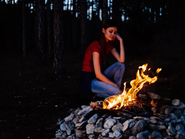 A woman sits near the fire and looks at the fire in a pine forest