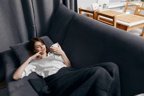 女人躺在沙发上，手里拿着电话，腿上挂着灰色的格子花。 — 图库照片