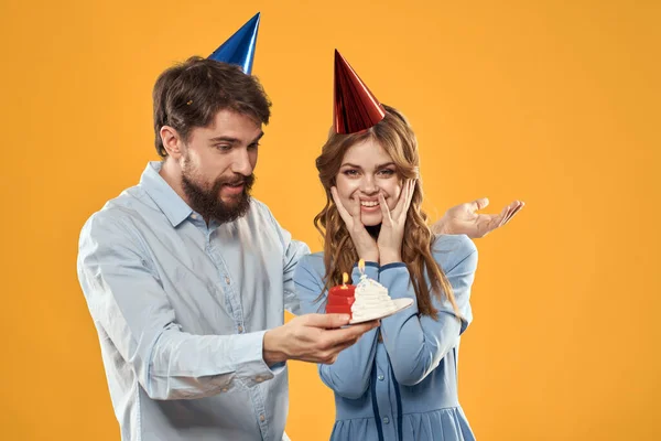Aniversário festa homem e mulher em um fundo amarelo em chapéus com um bolo em suas mãos — Fotografia de Stock